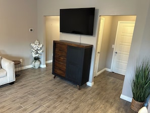 A living room with a flat-screen TV mounted above a dark wooden cabinet, a white sofa, and a small round side table. - Charming 3-bedroom house in peaceful neighborhood just 4 miles from OSU campus (Stillwater)