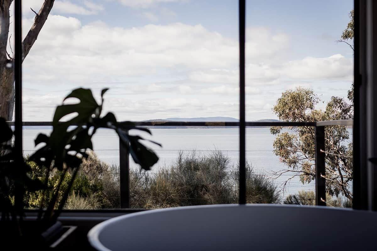 Une pièce avec vue sur une étendue d’eau et sur un paysage lointain, une fenêtre avec des stores verticaux et une plante en pot.