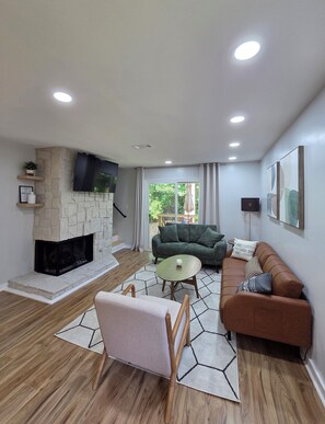 A living room with a stone fireplace, a green sofa, a brown sofa, a round table, and a TV mounted on the wall. - Stylish renovated 2BR - 5 min drive to Truist Park (Smyrna)