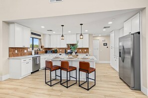 Una cocina moderna con armarios blancos, una isla central con asientos, electrodomésticos de acero inoxidable e iluminación empotrada en el techo.