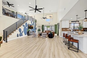 A spacious living area with a high ceiling, multiple ceiling fans, a staircase with a railing, and a kitchen area with a bar. - Casa Saguaro Old Town: 6BR Heated Pool Hot Tub (Scottsdale)