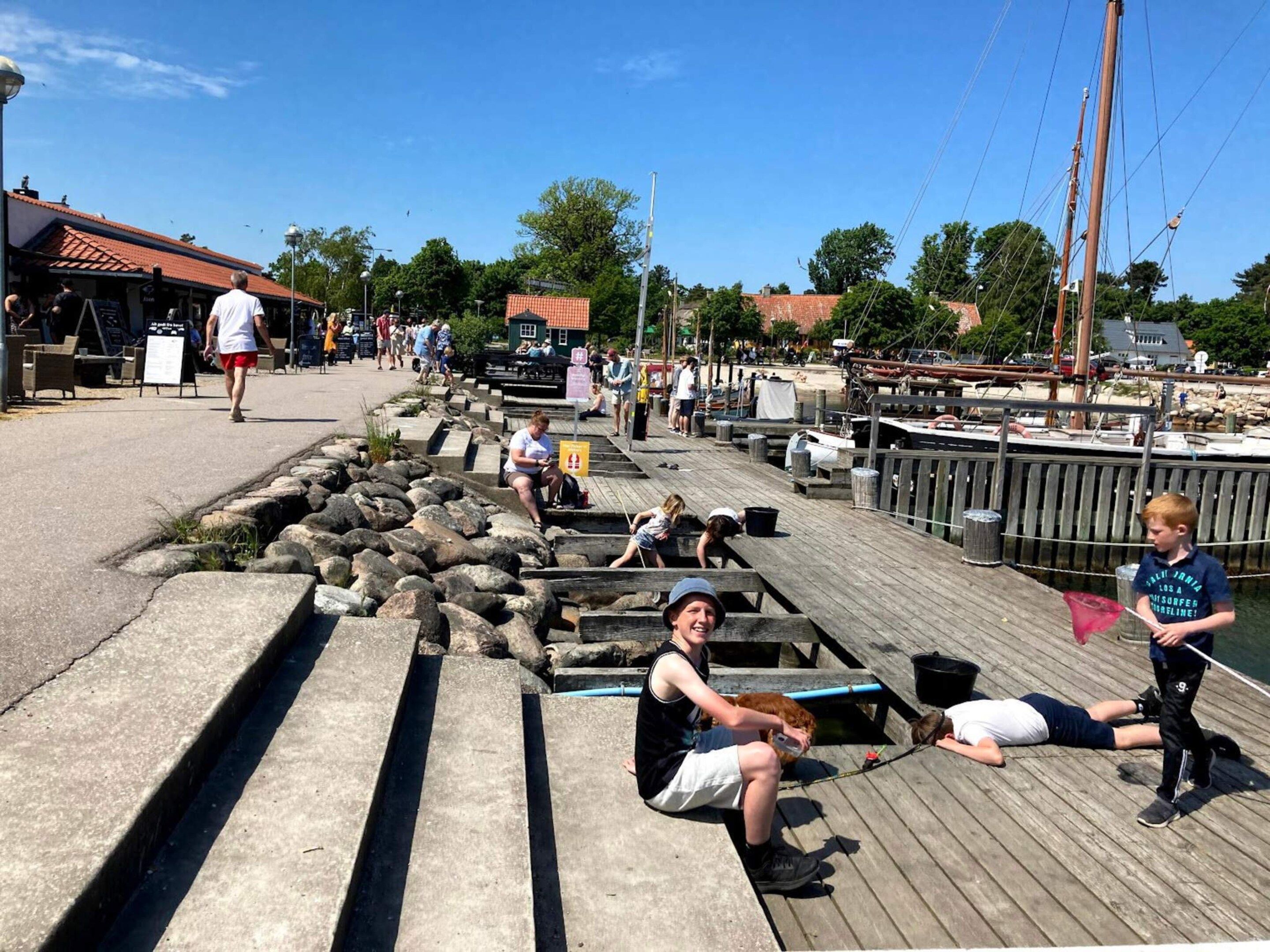 A wooden pier with people sitting and standing, a dock with boats, and a clear sky.