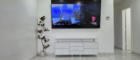 A modern living room with a flat-screen TV mounted on the wall, a white entertainment unit below, and a dark wood coffee table.