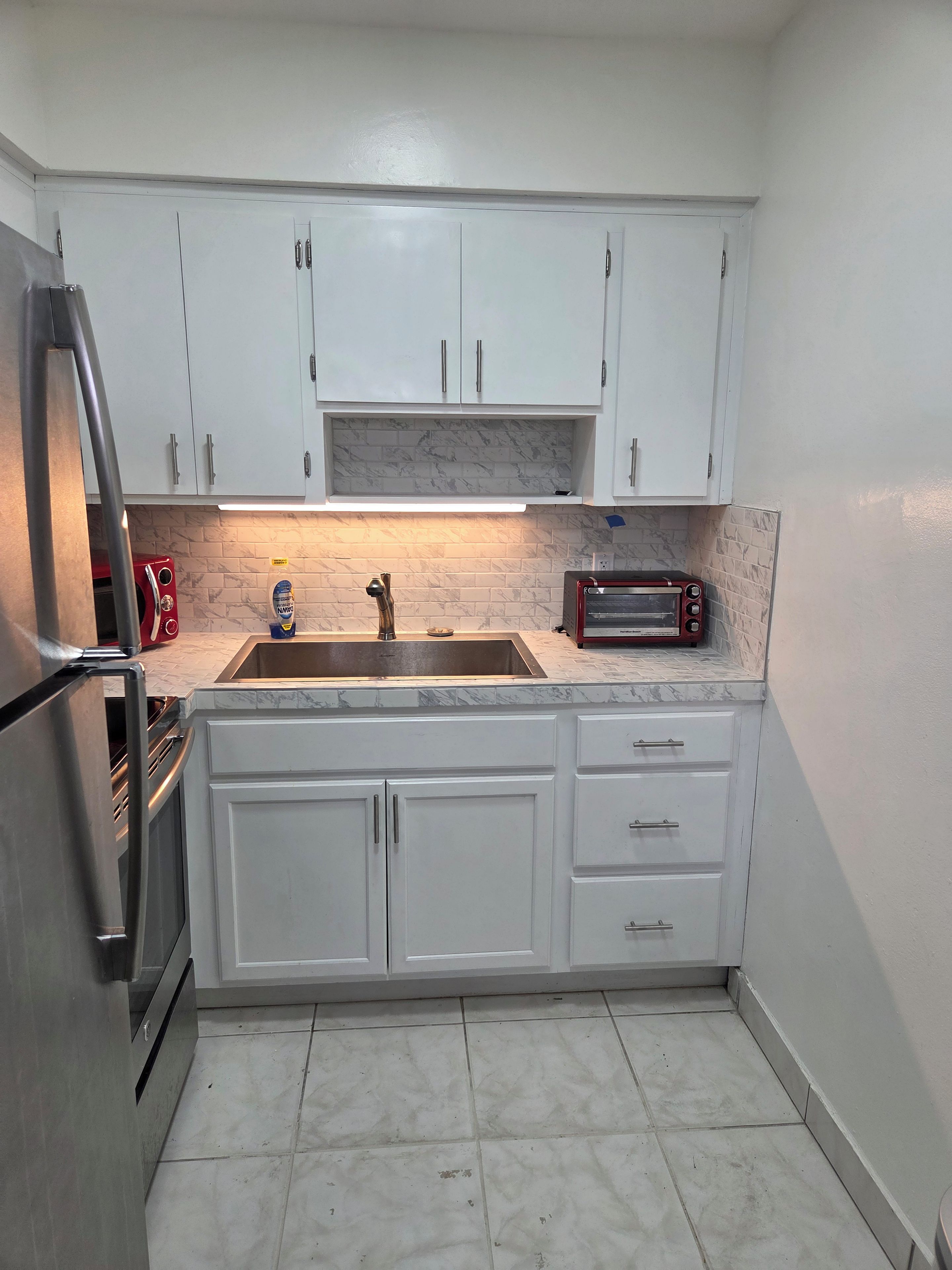 A compact kitchen with white cabinets, a stainless steel refrigerator, a built-in oven, a sink, and a red toaster.