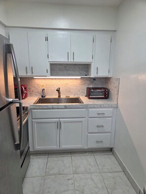 A compact kitchen with white cabinets, a stainless steel refrigerator, a built-in oven, a sink, and a red toaster. - Entire 2/1 Rental Unit in Fort Lauderdale (Lauderhill)
