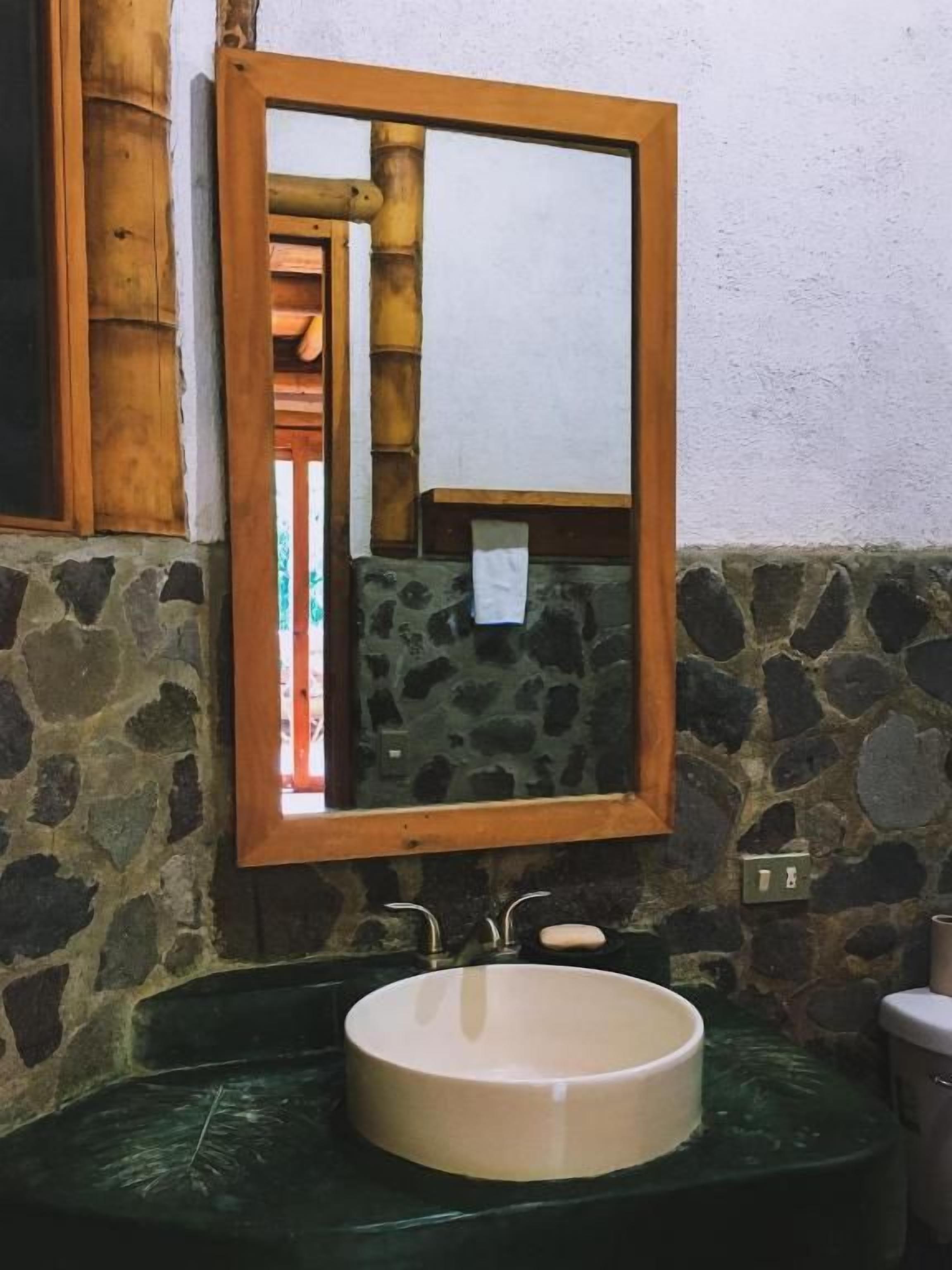 Um banheiro com parede de pedra, espelho com moldura de madeira, pia redonda e vaso sanitário.