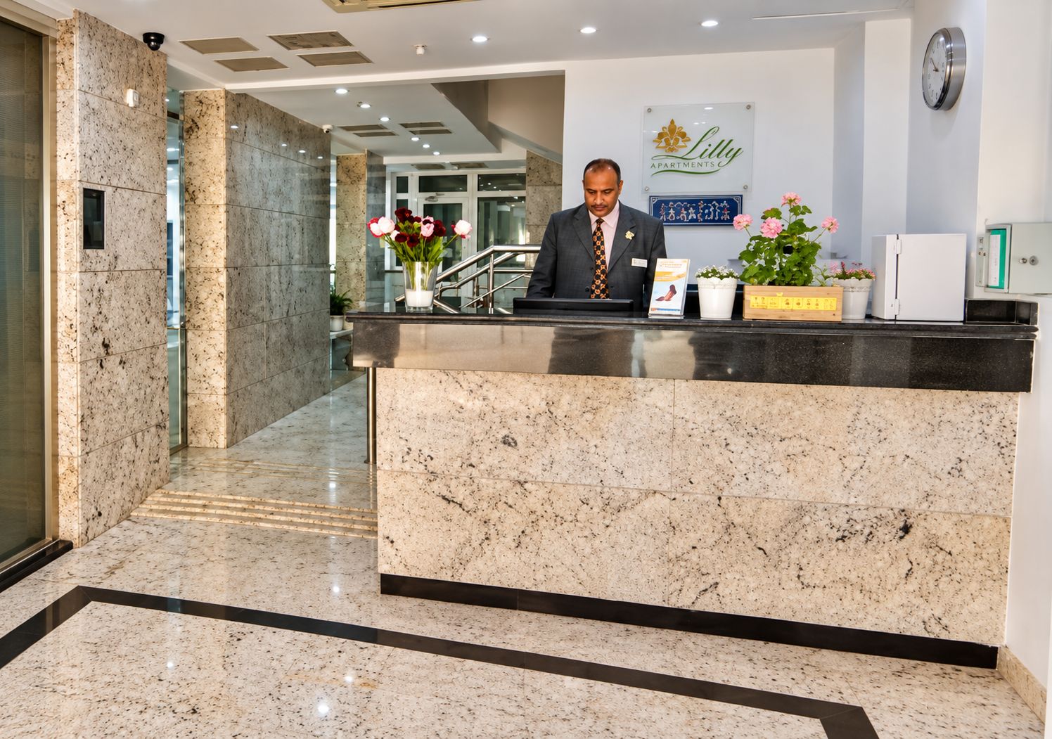 A hotel reception desk with a marble counter, a clock on the wall, and a sign for 'Lilly Apartments.'