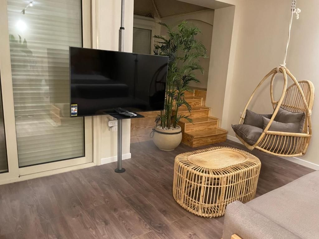 Una sala de estar con un televisor de pantalla plana, muebles de mimbre, una planta en maceta y una escalera al fondo.