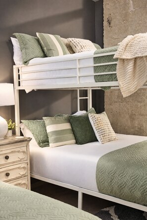 Ein Stockbett mit weißem Gestell, grün-weißer Bettwäsche und einem Nachttisch mit Lampe und Pflanze.