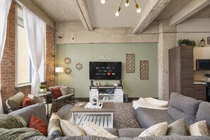 Ein Wohnzimmer mit einer Couch, einem Couchtisch und einem Fernseher. Im Hintergrund ist eine Ziegelwand und ein Küchenbereich zu sehen.