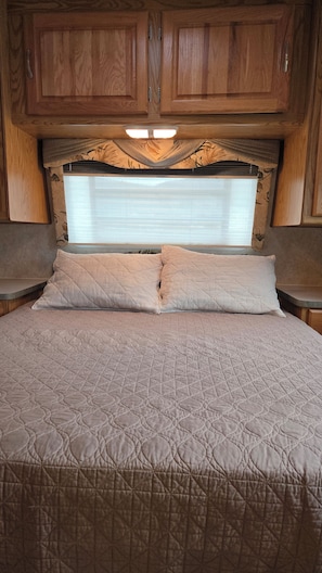 A bed with a quilted cover, wooden cabinets above, and a window with blinds.
