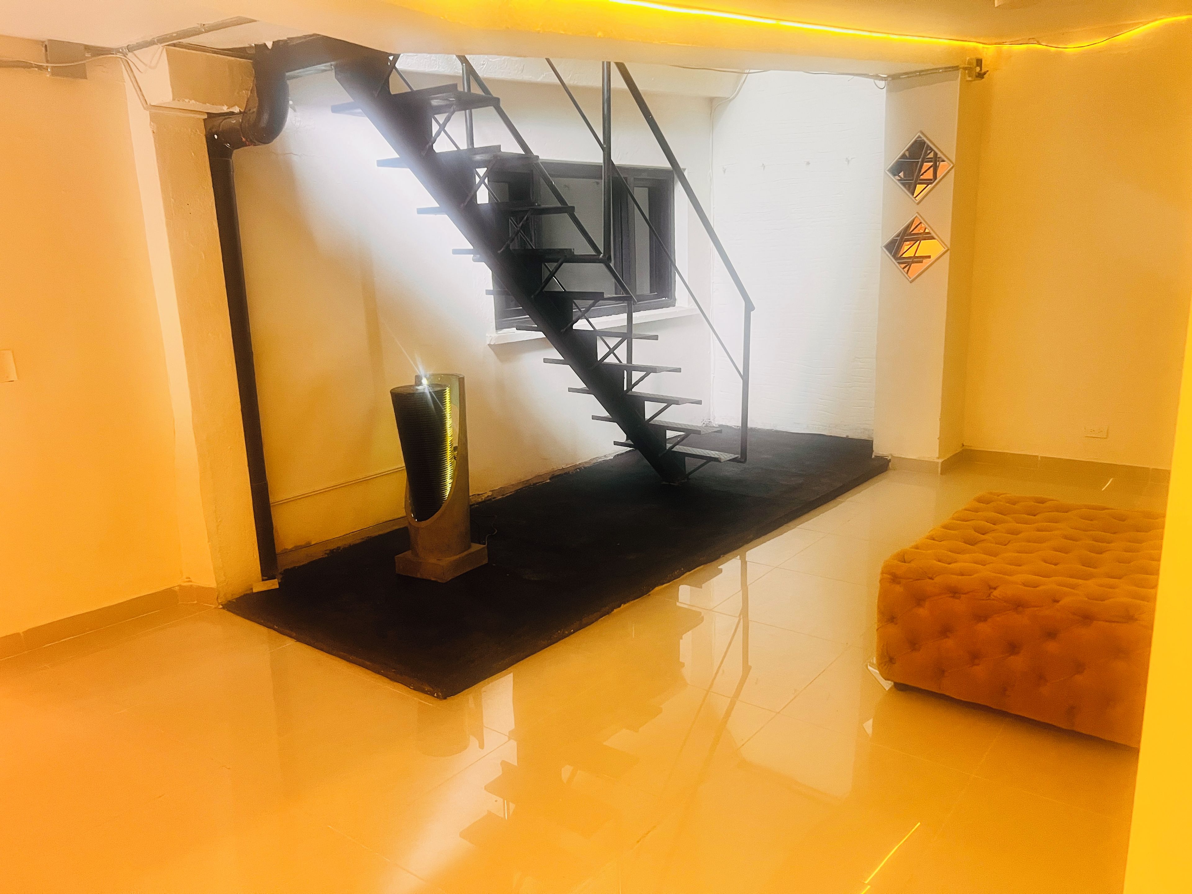 A modern interior with a staircase, a black mat, a tufted ottoman, and a metallic sculpture.