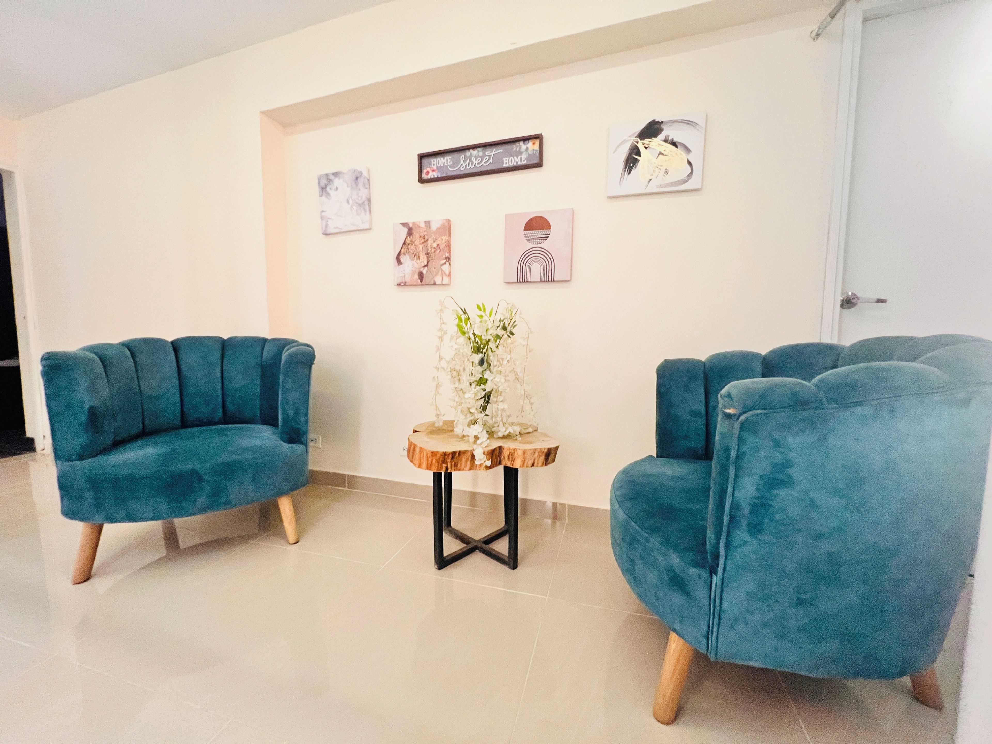 Una habitación con dos sillones azul turquesa, una mesita de madera con un jarrón de flores blancas y una puerta blanca.