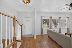 Un amplio espacio habitable con una escalera de madera, una puerta blanca y un ventanal con cortinas.