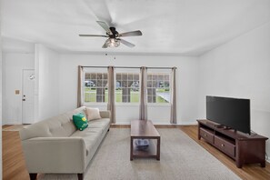 Een woonkamer met een plafondventilator, een flatscreen-tv, een bank, een salontafel en een raam met gordijnen.