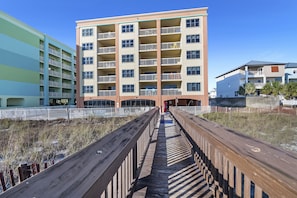 A multi-story building with balconies, a wooden boardwalk leading to the entrance, and a clear blue sky. - Harbour Place 210 | Beachfront | Pools | Heart of Orange Beach (Orange Beach)