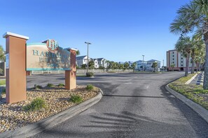 Un panneau indiquant l’accès à « Harbour Place », avec une route bien dégagée menant à une entrée fermée par un portail, le tout entouré de palmiers et d’une clôture.