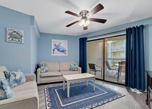 A living room with a ceiling fan, a sofa, a coffee table, and a patterned rug. - Harbour Place 210 | Beachfront | Pools | Heart of Orange Beach (Orange Beach)