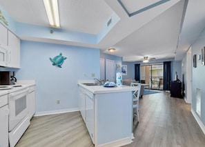 A modern kitchen with a central island, a dining area with a table and chairs, and a living space with a sofa and a TV. - Harbour Place 210 | Beachfront | Pools | Heart of Orange Beach (Orange Beach)