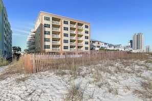 A multi-story hotel with balconies, located near a sandy beach area with a wooden fence. - Harbour Place 210 | Beachfront | Pools | Heart of Orange Beach (Orange Beach)
