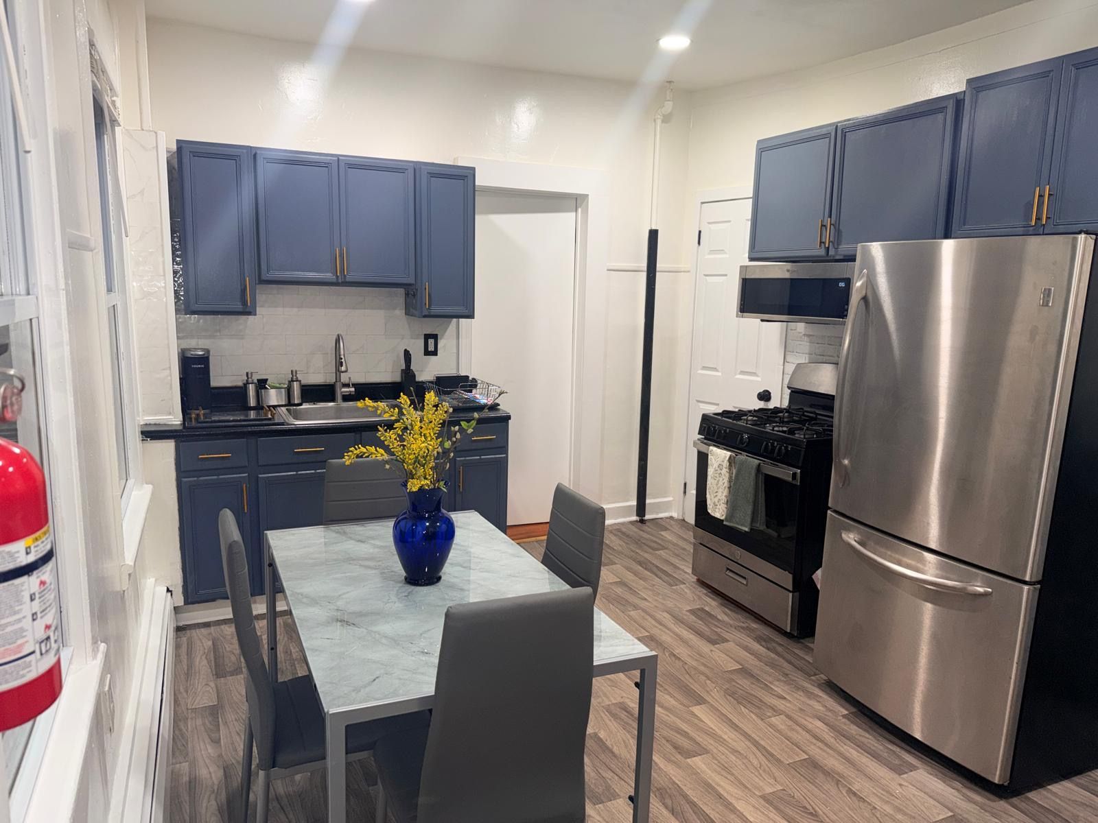 A compact kitchen with dark blue cabinets, a stainless steel refrigerator, a stove, and a marble countertop with a vase of flowers.