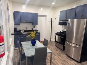 A compact kitchen with dark blue cabinets, a stainless steel refrigerator, a stove, and a marble countertop with a vase of flowers.