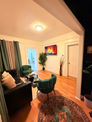 A living room with a sofa, a green armchair, a coffee table, and a rug. There is a painting of a fire on the wall.