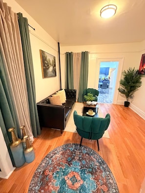 Un salon comprenant un canapé, un fauteuil vert, un tapis à motifs et un coin repas visible à travers une porte ouverte.