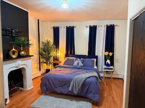 A bedroom with a bed, a nightstand, a lamp, a fireplace, and curtains.