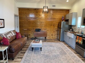 A cozy living room with a wooden accent wall, a brown leather sofa, a coffee table, and a kitchen area with a refrigerator and cabinets. - Delightful 1-Bedroom Barn Apartment Located In Gated Equestrian Community (Aiken)