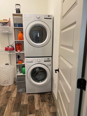 A laundry room with two stacked front-loading washing machines, a shelf with various laundry supplies, and a trash bin. - Delightful 1-Bedroom Barn Apartment Located In Gated Equestrian Community (Aiken)