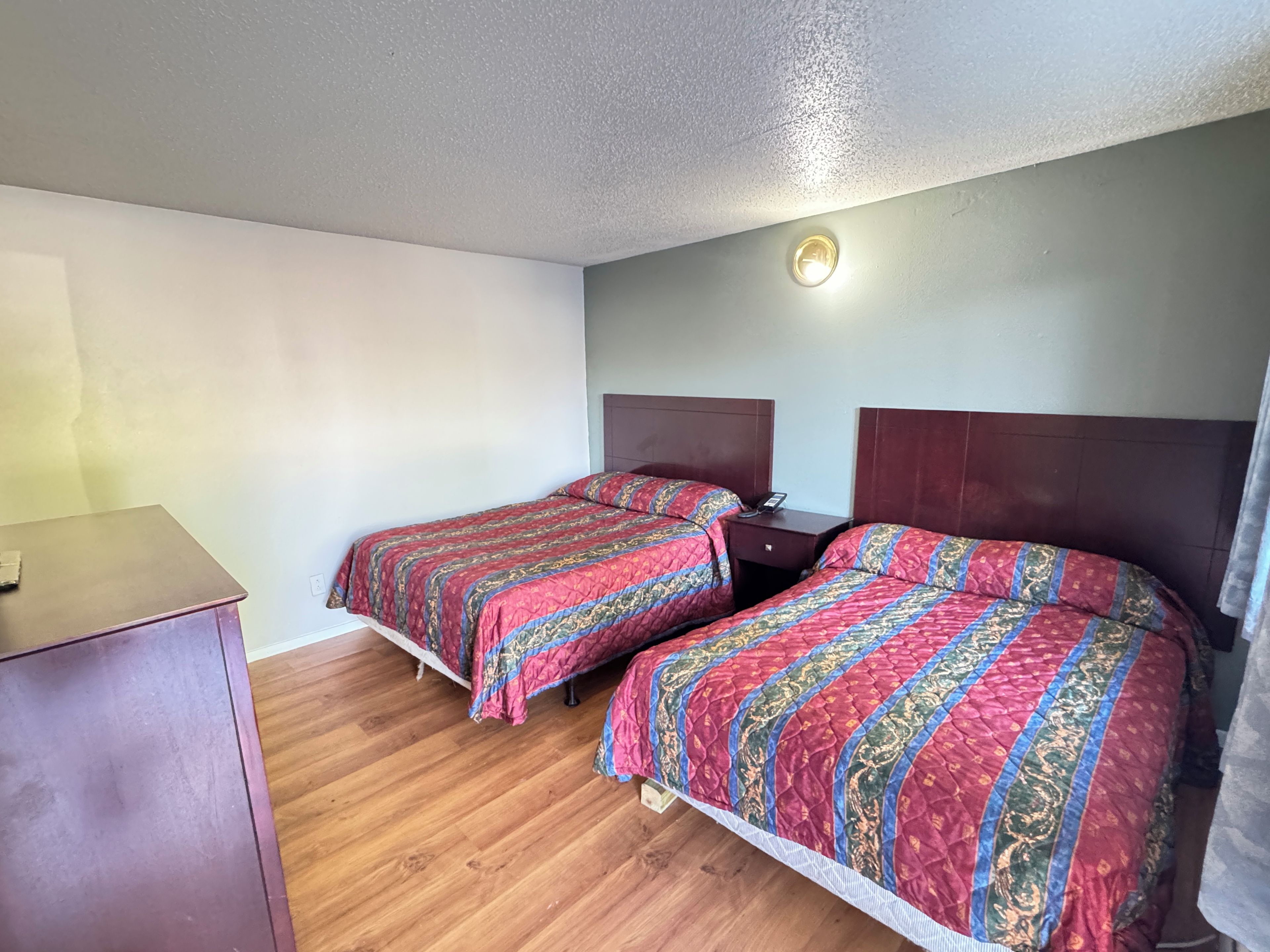A room with two beds, a wooden floor, a desk, and a wall-mounted light fixture.