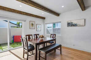 Un coin repas avec une table en bois, des chaises et un banc. Il y a deux fenêtres avec des cadres blancs et un tableau au mur.