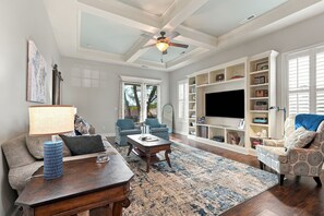 Uma sala de estar ampla com ventilador de teto, uma grande TV de tela plana, uma estante de livros e um espaço de estar confortável com mesa de centro e sofás.
