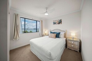 A bedroom with a large bed, a window with curtains, a ceiling fan, and a nightstand with a lamp. - Coastal Outlook - Caloundra Poolside Apartment (Caloundra Region)