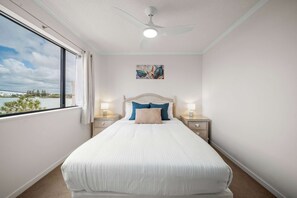 A bedroom with a large bed, two bedside tables with lamps, a ceiling fan, a window with a view, and a wall-mounted artwork. - Coastal Outlook - Caloundra Poolside Apartment (Caloundra Region)