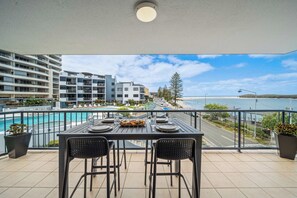 A balcony with a table set for two, overlooking a swimming pool and a beachfront. - Coastal Outlook - Caloundra Poolside Apartment (Caloundra Region)