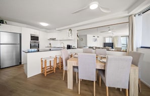 A modern kitchen with a built-in oven and refrigerator, a dining area with a wooden table and chairs, and a large window overlooking a body of water. - Coastal Outlook - Caloundra Poolside Apartment (Caloundra Region)