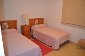 A small, simple room with two beds, a nightstand, a lamp, and a window with a blind. - Bahía Meloneras Villas Bahmel01 (San Bartolomé de Tirajana)