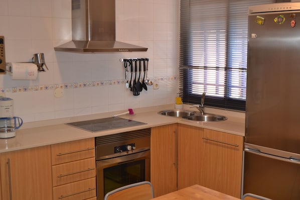 A kitchen with a stainless steel range hood, a built-in oven, a refrigerator, and a window with blinds. - Bahía Meloneras Villas Bahmel01 (San Bartolomé de Tirajana)
