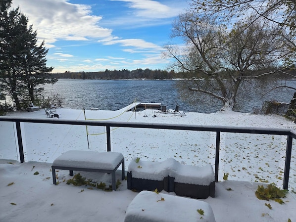 A snow-covered deck with a bench and a table, overlooking a lake with a dock and trees.