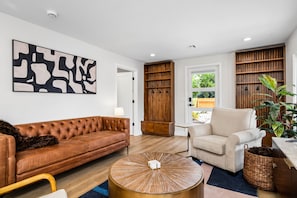 A modern living room with a brown leather sofa, a white armchair, a round coffee table, and a large abstract painting on the wall. - South Campus Modern - Upscale & Walkable to UO, Park & Shops (Eugene)