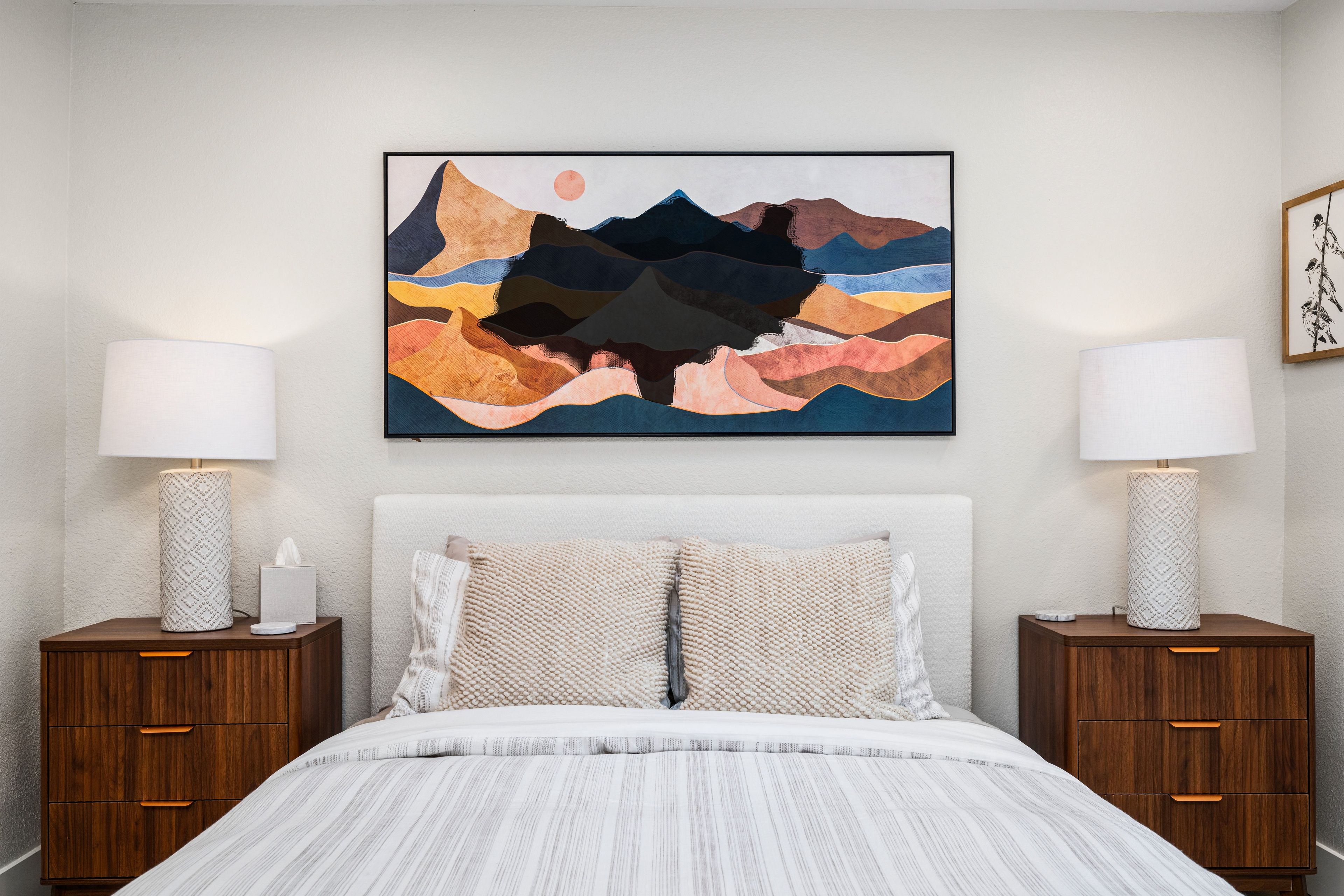A bedroom with a bed, two bedside tables, a wall-mounted abstract painting, and a wall-mounted lamp.