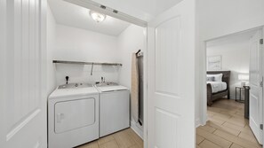 A laundry room with two white washing machines, a ceiling light, and a door leading to a bedroom with a bed, nightstand, and lamp.