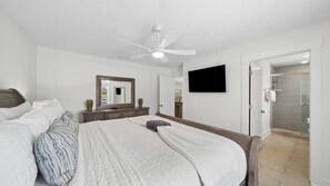 Ein Schlafzimmer mit einem Bett, einem Schminktisch, einem Spiegel, einem Deckenventilator, einem Fernseher und einer Dusche.