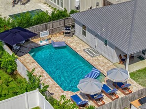 Ein rechteckiger Swimmingpool mit einer gefliesten Liegefläche, umgeben von einem Holzzaun. Liegestühle und Sonnenschirme sind vorhanden, und ein Grill ist sichtbar.