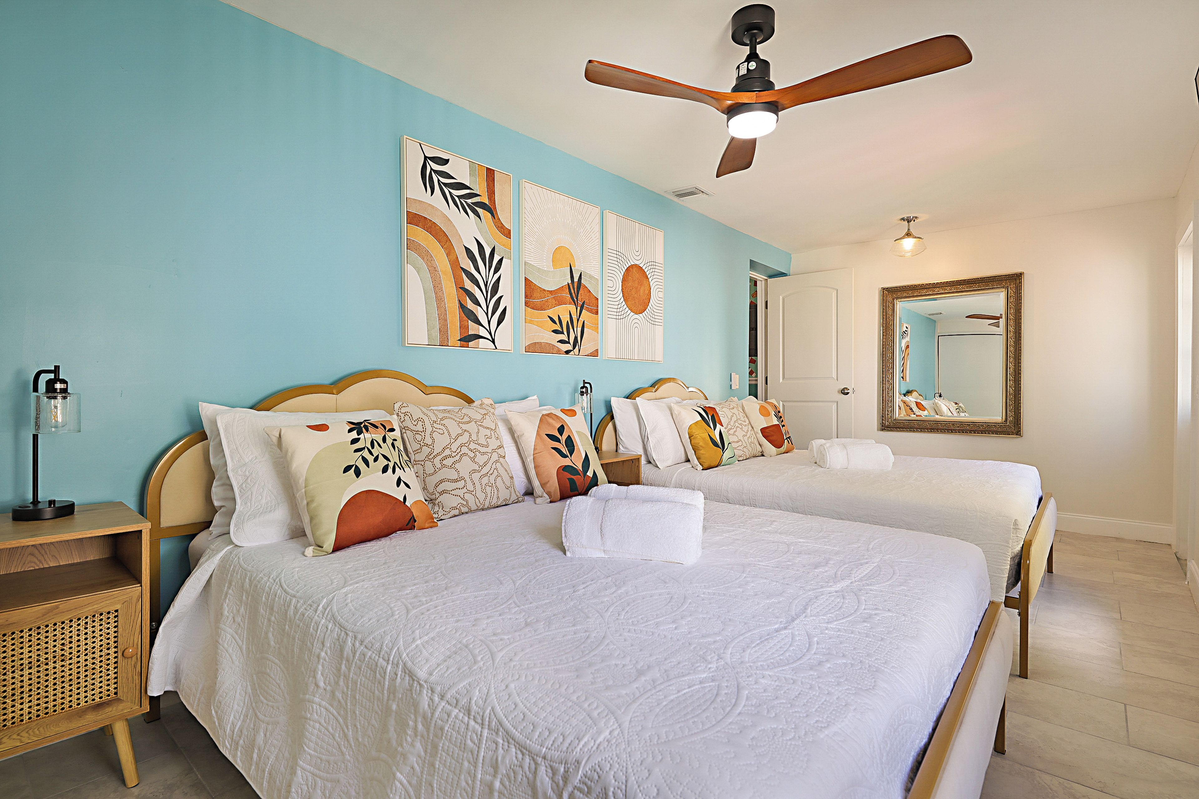 A hotel room with two beds, a ceiling fan, artwork on the wall, and a mirror.