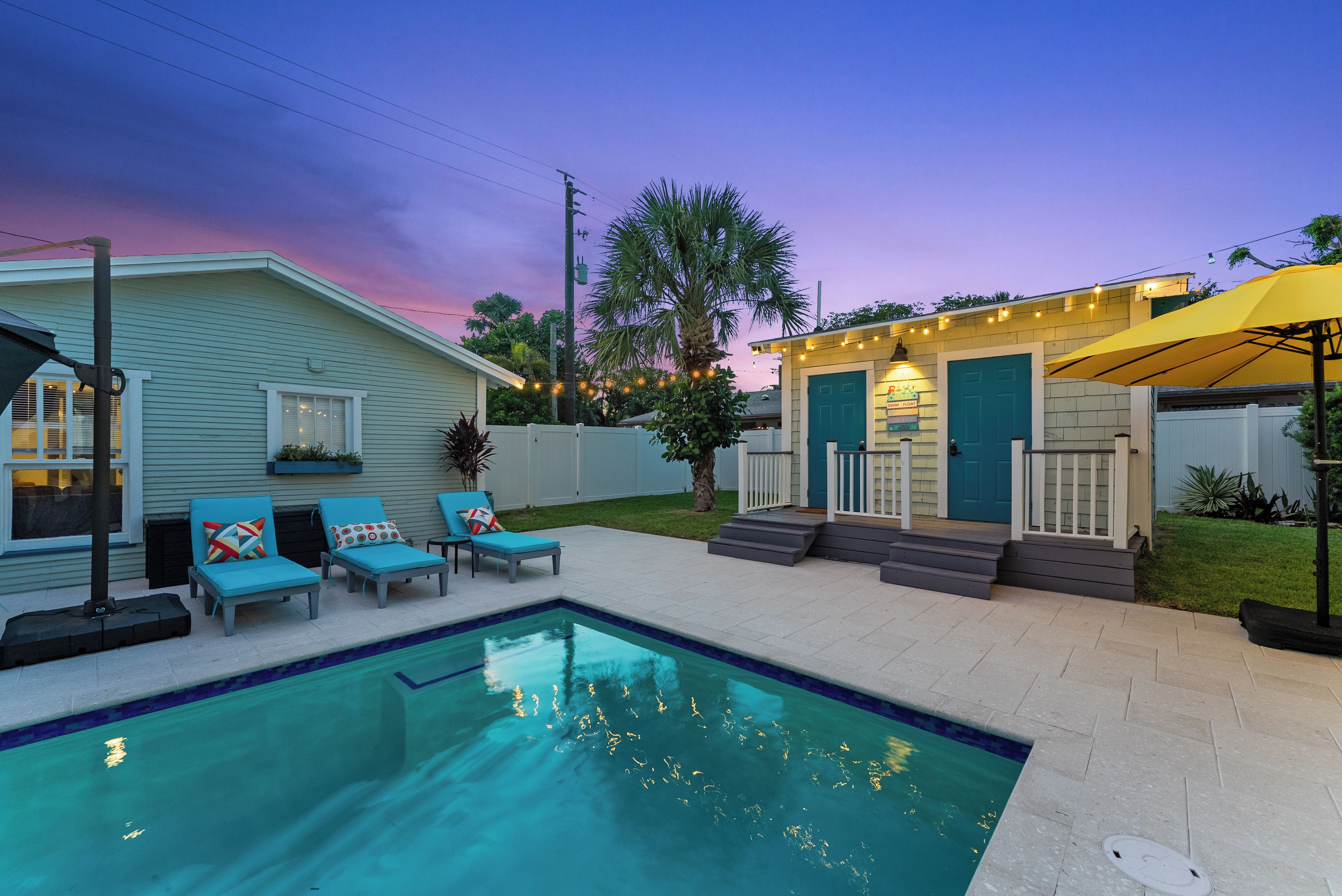 A backyard with a swimming pool, lounge chairs, a small deck, and a house with a blue door.
