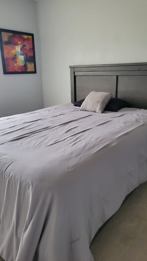 Ein ordentlich bezogenes Bett mit grauem Kopfteil und einem Kissen, ein gerahmtes abstraktes Gemälde an der Wand und ein Teppichboden.
