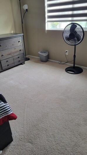 A room with a beige carpet, a dresser with drawers, a standing fan, a trash can, and a window with blinds. - Spacious 4-bedroom home in pleasant Moreno Valley ,nearby golf and lake nearby (Moreno Valley)